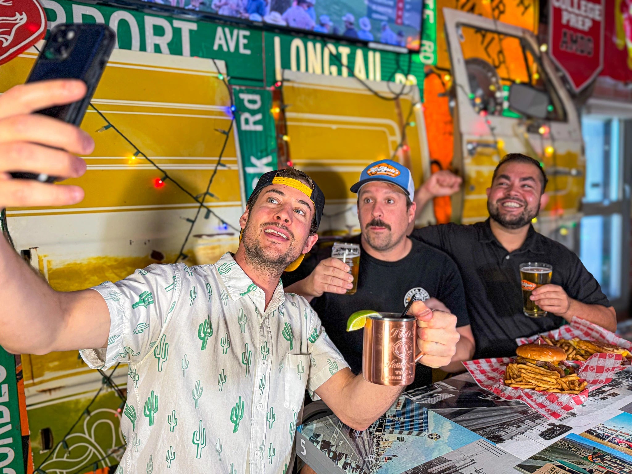 Three men taking a selfie in a bar with drinks and snacks on a table.